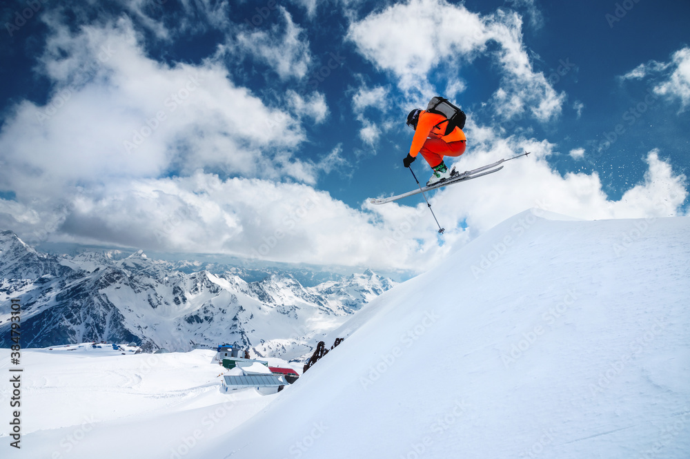 Man skier athlete makes a jump in flight on a snowy slope against the backdrop of a blue sky of mountains and clouds. Freeride and extreme skiing for men Man skier athlete makes a jump in flight on a snowy slope against the backdrop of a blue sky of mountains and clouds. Freeride and extreme skiing for men
