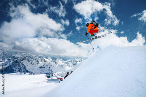 Man skier athlete makes a jump in flight on a snowy slope against the backdrop of a blue sky of mountains and clouds. Freeride and extreme skiing for men