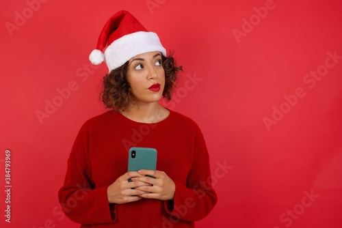Portrait of beautiful young woman wearing Christmas hat with dreamy look  thinking while holding smartphone. Tries to write up a message for her father  explaining why she took car