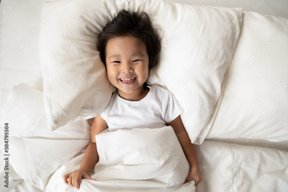 Top view portrait smiling cute Asian little girl lying in cozy bed, laughing pretty toddler child kid looking at camera, resting on soft pillow under warm duvet blanket, enjoying lazy leisure time Top view portrait smiling cute Asian little girl lying in cozy bed, laughing pretty toddler child kid looking at camera, resting on soft pillow under warm duvet blanket, enjoying lazy leisure time