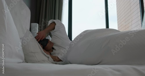 Pretty young woman awakes and takes off sleep mask lying on large bed with pillows near window in hotel room in morning