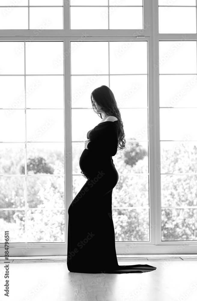 beautiful young pregnant woman in dark black dress stands near the window black and white photo beautiful young pregnant woman in dark black dress stands near the window black and white photo