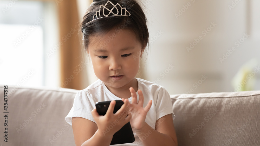 Close up cute little girl wearing princess diadem using smartphone, pretty toddler child looking at screen, playing mobile device game, watching cartoons online, sitting on cozy couch at home Close up cute little girl wearing princess diadem using smartphone, pretty toddler child looking at screen, playing mobile device game, watching cartoons online, sitting on cozy couch at home