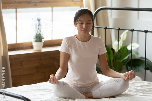 Calm peaceful Asian young woman meditating in bedroom alone, tranquil beautiful girl with closed eyes sitting in lotus pose on cozy bed, dreaming, doing yoga exercise, stress relief concept
