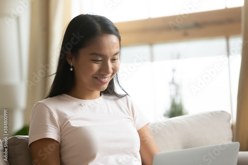 Free Download Close up smiling young Asian woman looking at laptop screen, watching funny video, movie, chatting in social network with friends, shopping or studying online, sitting on cozy couch at home - GFXLoad
