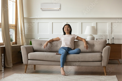 Tranquil calm young Asian woman relaxing in room with air conditioner on wall, sitting on cozy couch, smiling peaceful girl resting on sofa in living room, enjoying fresh air, breathing