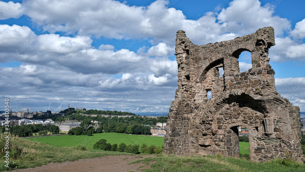 Summer photo of Saint Antony's Chapel ruins in Holyrood Park. With Edinburgh Skyline and famous landmarks in the background, including Calton Hill and the city's palace and gardens in Scotland UK