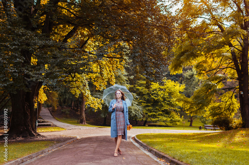 Stylish young woman walking in park with transparent umbrella wearing trendy outfit. Autumn fashion