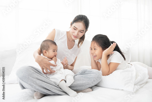 Asian mother and children girl baby laugh together at home. Asia mom holdind infant and play with sister sitting on white bed in living room at house family having fun together.