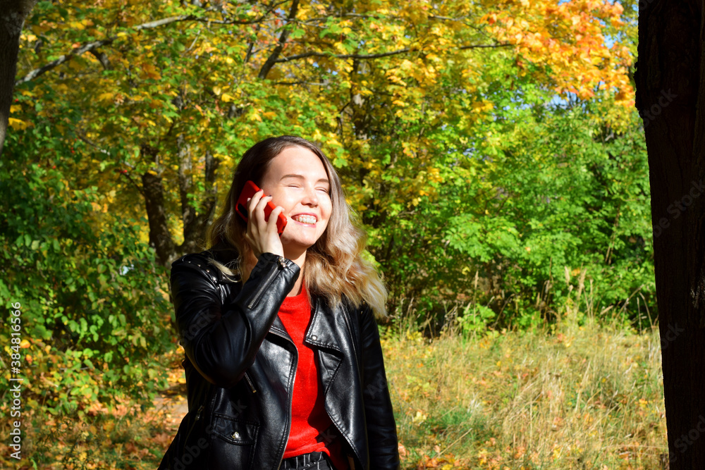 Young woman is smiling and talking on the phone in the autumn park on a sunny day. Concept of distance communications Young woman is smiling and talking on the phone in the autumn park on a sunny day. Concept of distance communications