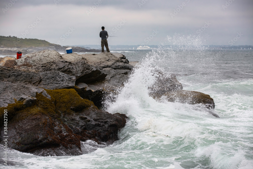 A fisherman with fishing equipment, fishing on top of dangerous rocky coastline with waves crashing in Beavertail State Park, Rhode Island