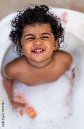 Image depicting Baby hygiene having a Cheerful Indian toddler bathing in bath tub. Baby girl enjoying her bath with toys and soapy foam.