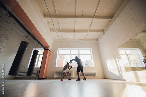 A man with boxing paws on his hands teaches the technique of hitting a novice girl in a light gym