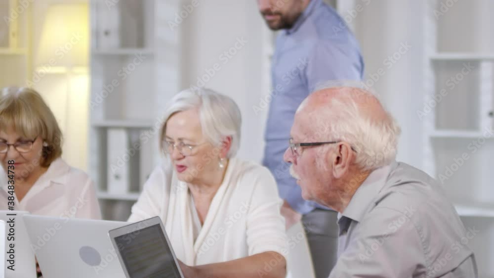Tracking shot of male technology teacher with beard looking over shoulders of elderly people using laptops and giving them advice during computer class Tracking shot of male technology teacher with beard looking over shoulders of elderly people using laptops and giving them advice during computer class