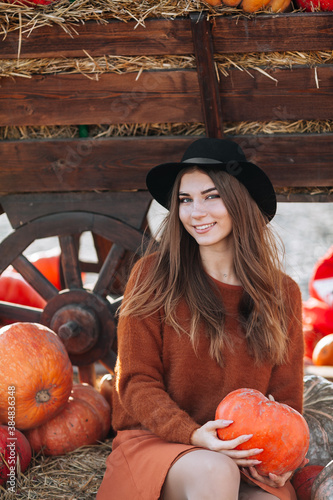 Portrait of happy smiling woman sitting near wooden wagon with orange pumpkin on farmers market in brown sweater, dress and hat. Cozy autumn vibes Halloween, Thanksgiving day