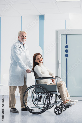 Senor doctor standing near patient in wheelchair in clinic