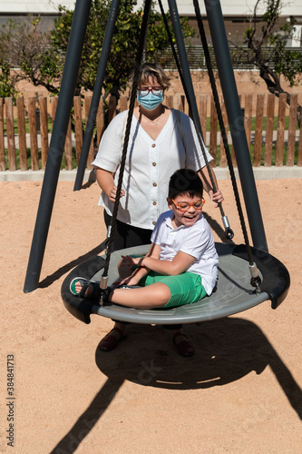 disabled child in swing set with mother wearing a pandemic coronavirus protective mask