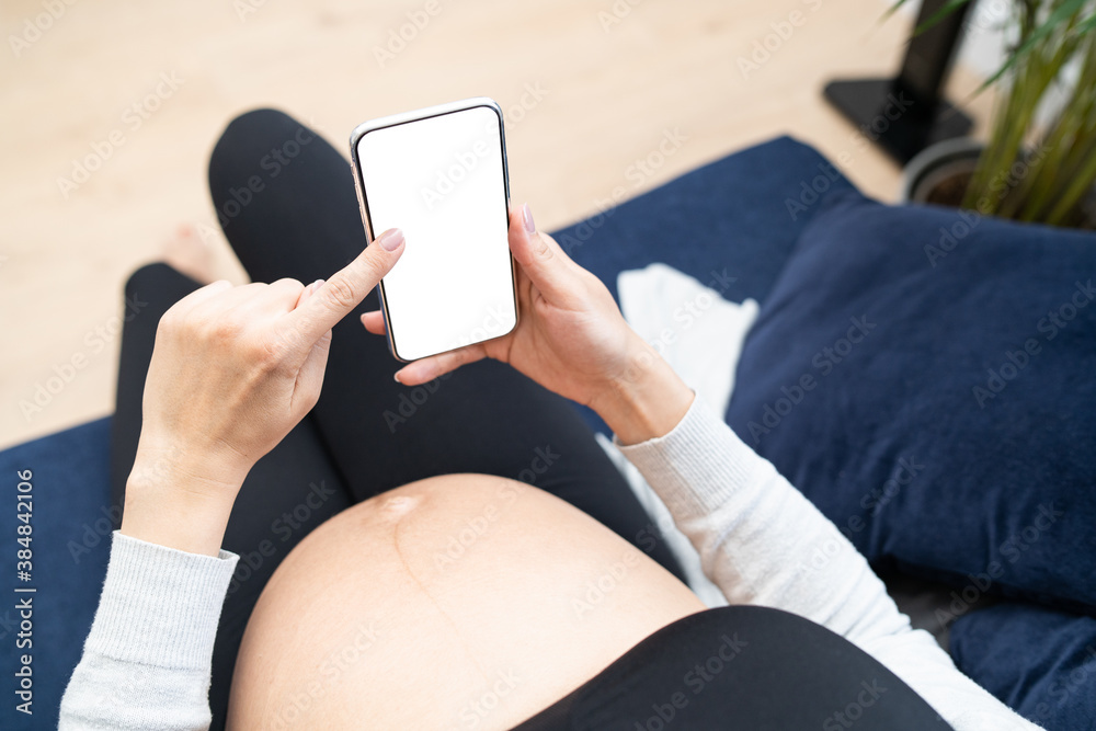 Young pregnant female using cell phone during relax in living room. Caucasian young mom has on online conversation with family. Top view of pregnant belly.