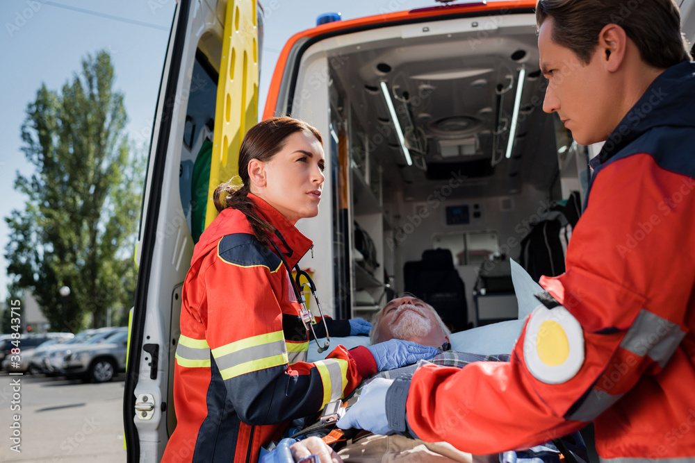 Selective focus of paramedics in uniform standing near senior patient on stretcher and ambulance auto on urban street Selective focus of paramedics in uniform standing near senior patient on stretcher and ambulance auto on urban street
