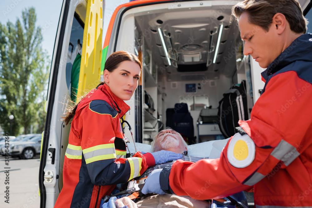 Selective focus of paramedic looking at camera near patient on stretcher and colleague outdoors Selective focus of paramedic looking at camera near patient on stretcher and colleague outdoors