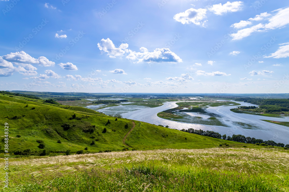 Sunny summer landscape with river,fields,green hills and beautiful clouds in blue sky.Pretty view of summer scenery on a nice day.Green trees growing along the river.