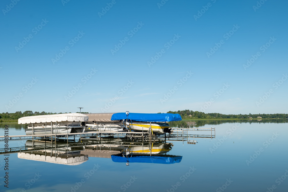 Docked boats on lifts are reflected in the calm water of a pretty lake in Minnesota, on a sunny summer day with beautiful clear blue sky.