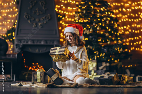 Pretty woman in warm sweater, socks and christmas hat, sitting on the floor at home with gift boxes