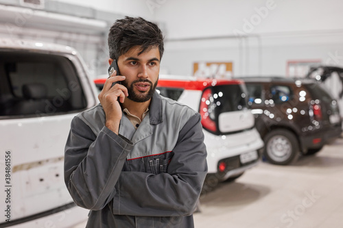 serious auto mechanic man talk on phone with boss, stand looking away, in uniform. discuss something