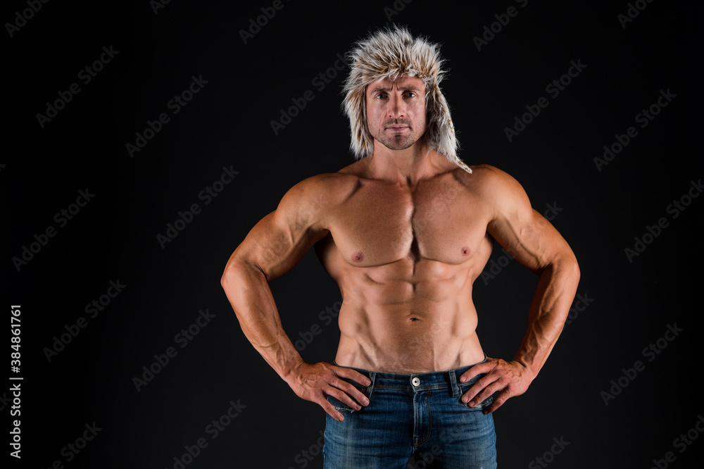 Naklejka premium Bodycare with fitness and sport. Man with muscular torso. Bodybuilder in fur hat and jeans dark background. Bodycare and wellness. Health and diet