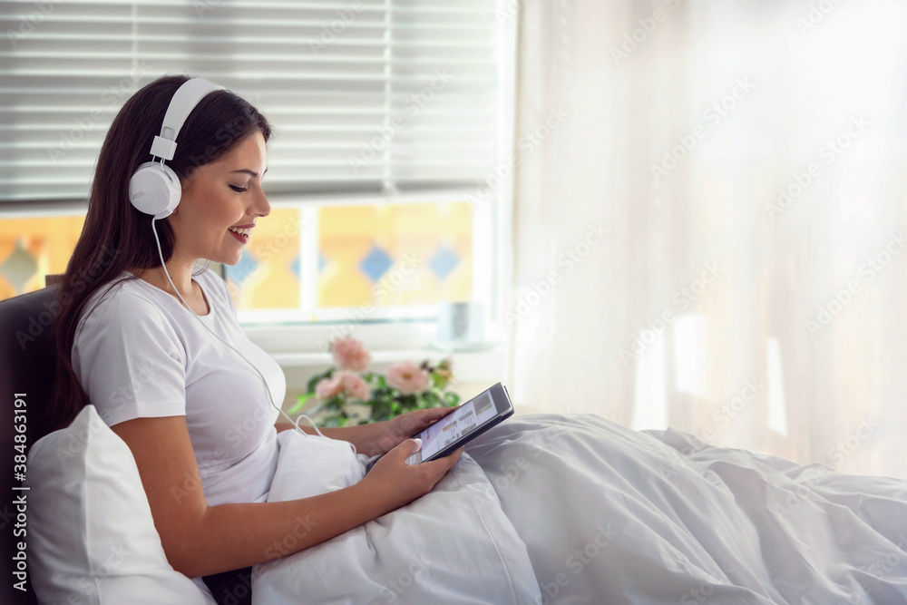 A beautiful, young woman wearing headphones and reading the morning news on her tablet device in bed wilst listening to music A beautiful, young woman wearing headphones and reading the morning news on her tablet device in bed wilst listening to music