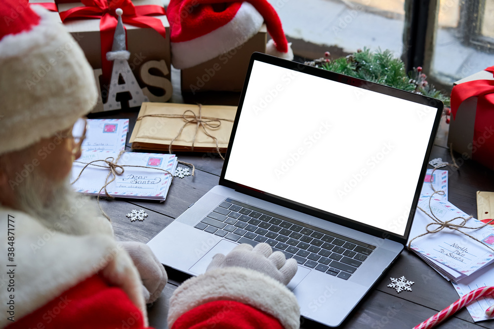 Over shoulder view of Santa Claus wearing costume using laptop computer with white blank empty mock up screen monitor sitting at workshop table on Merry Christmas eve. E commerce website ads concept - obrazy, fototapety, plakaty 