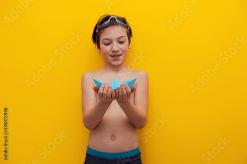 A cheerful boy of 9 years old examines a paper boat. Photo of a cheerful boy who is going to swim on a yellow background. Studio photo. boy tourist in swimming trunks