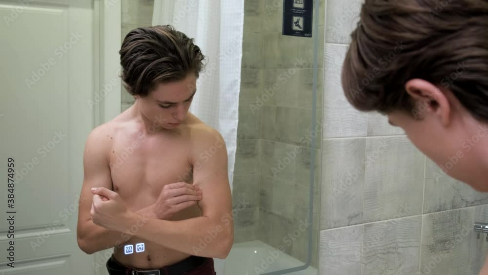 A teenager looks at his body in the mirror. The guy dreams of a muscular body