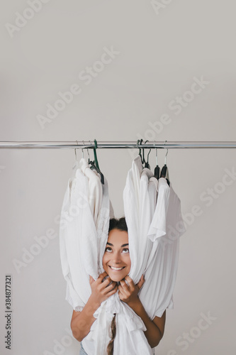 Beautiful young woman stylist standing near rack with hangers with white clothes. Shopaholic with many clothes..