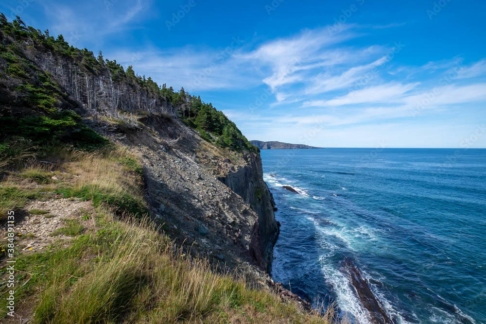 A hiking trail along the ocean with waves crashing on the jagged rocks. The calm water and sky are blue with white clouds. There are trees, rocks, grass and cliff along the edge of the Atlantic ocean. A hiking trail along the ocean with waves crashing on the jagged rocks. The calm water and sky are blue with white clouds. There are trees, rocks, grass and cliff along the edge of the Atlantic ocean.