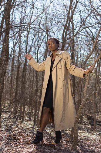 Photo of young African American woman walking through bare winter forest. Focus on face. Black shirt dress and creme color winter coat. Hands on tree branches.