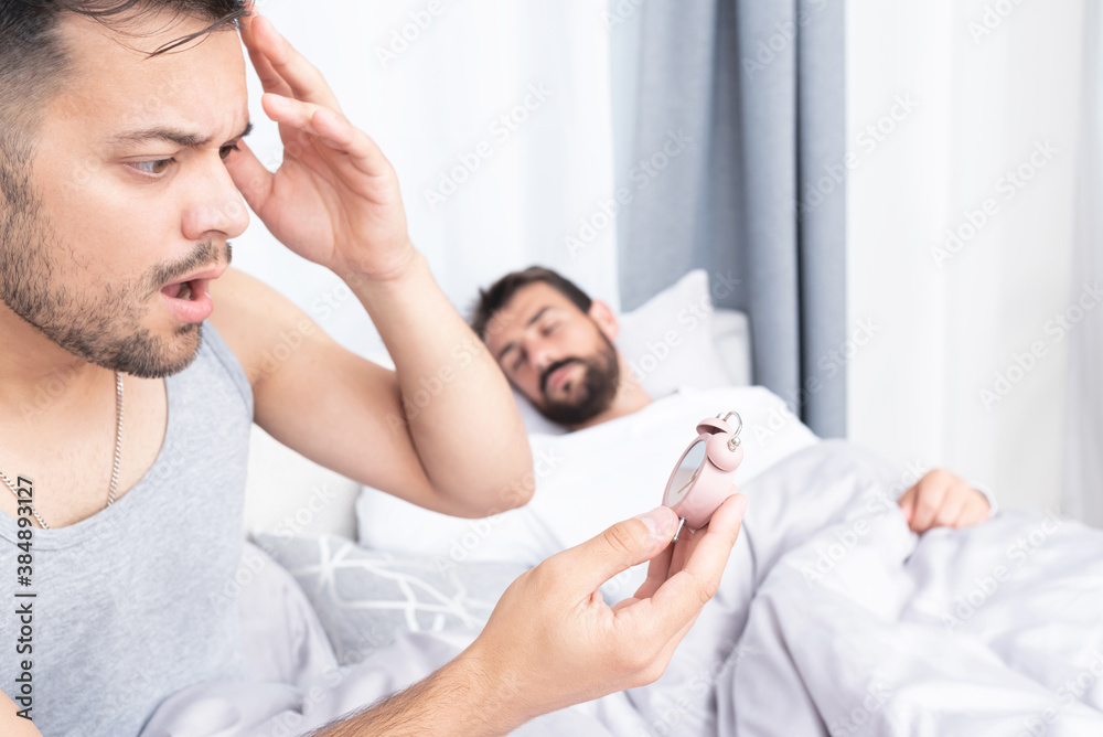 Worried young man being late for work with boyfriend sleeping in bed Worried young man being late for work with boyfriend sleeping in bed