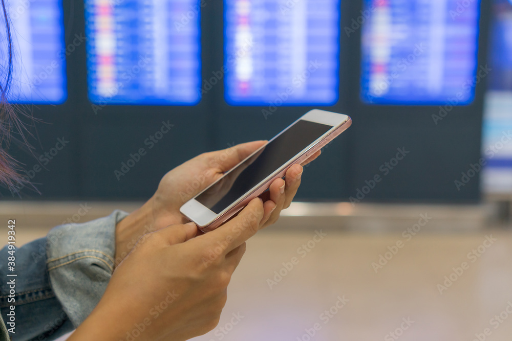 Hands of young Asian tourist woman using smartphone near flight information board in international airport