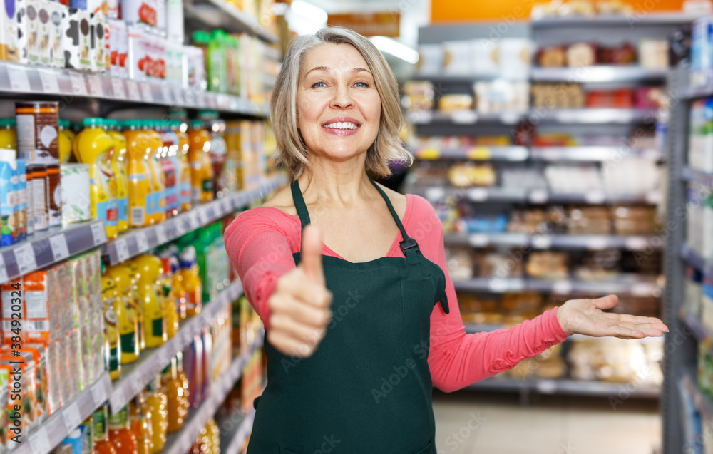 Smiling confident mature saleswoman giving thumbs up, recommending quality food products in the supermarket