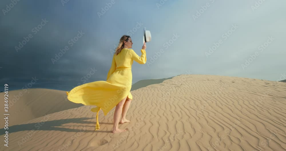 Cinematic slow motion of woman walking by sand dune. Barefoot female traveler in waving on the wind dress by the rippled sand desert surface with dark grey cloud on background. 4K Scenic nature 