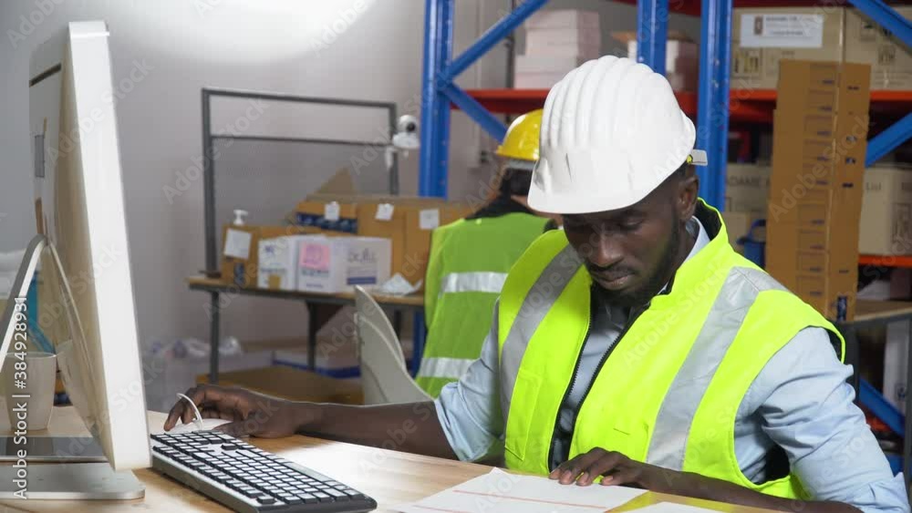 african warehouse manager sitting at desk using computer check inventory items online at distribution warehouse. black man worker in Hard Hat typing keyboard entering data document in workplace african warehouse manager sitting at desk using computer check inventory items online at distribution warehouse. black man worker in Hard Hat typing keyboard entering data document in workplace