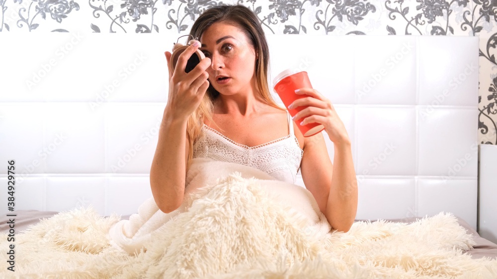 Attractive young woman gets up in bed with a cup of coffee and an alarm clock. The woman overslept and is late, reacting with horror at the time. Attractive young woman gets up in bed with a cup of coffee and an alarm clock. The woman overslept and is late, reacting with horror at the time.