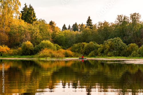 View of the calm water of the lake with the reflection of the autumn forest. A rubber rowing boat with a fisherman and a dog is visible in the distance