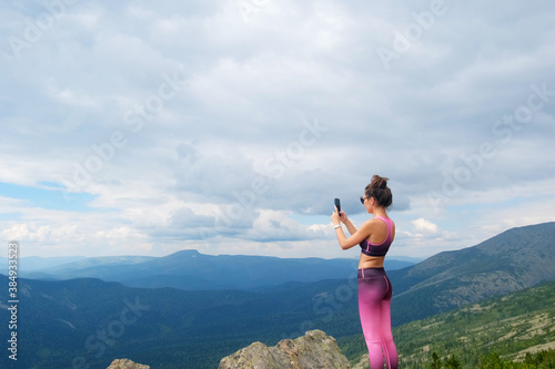 A woman take a photo on smartphione on top of rock in mountains. Young woman hiking up hill against a blue sky with clouds.