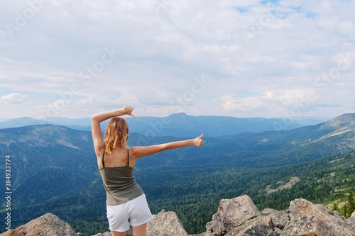 A woman jumps, dances, and climbs, does yoga on top of mountains. Young woman hiking up hill against a blue sky with clouds. © evelinphoto