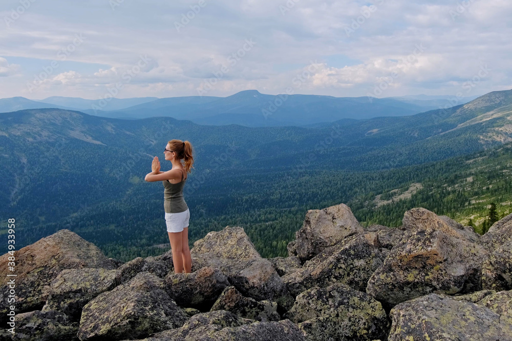 A woman doing yoga exercises in prayer posture on top of mountains. Young woman hiking up hill against a blue sky with clouds.