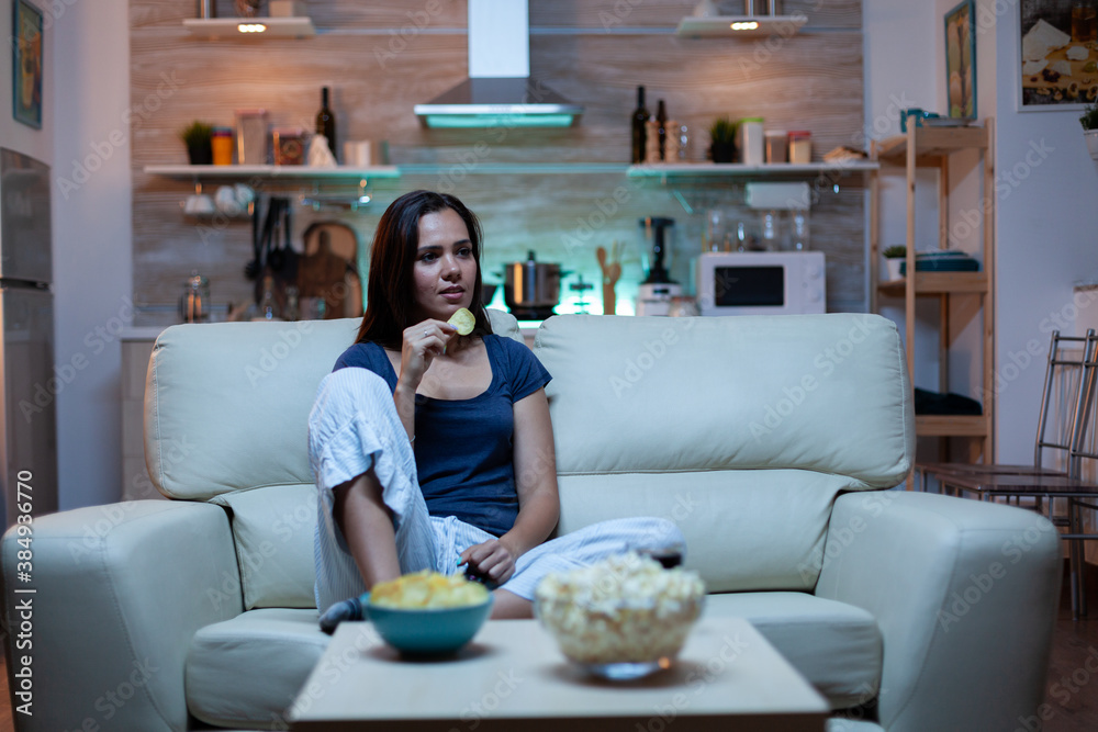 Happy woman eating popcorn on sofa and watching tv in living room at home. Excited, amused, lonely lady enjoying the evening sitting on comfortable couch dressed in pajamas in front of television.