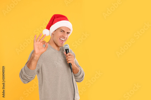 Emotional man in Santa Claus hat singing with microphone on yellow background, space for text. Christmas music © New Africa
