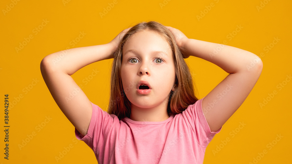 Confused child. Problem anxiety. Omg reaction. Stress disappointment. Portrait of overwhelmed shocked young girl clutching head looking at camera isolated on orange background. Confused child. Problem anxiety. Omg reaction. Stress disappointment. Portrait of overwhelmed shocked young girl clutching head looking at camera isolated on orange background.