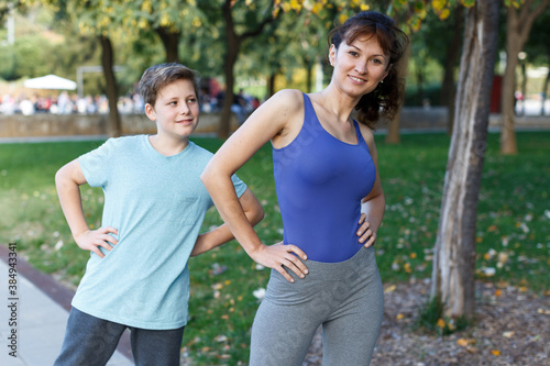 Smiling sporty woman and tweenager boy doing exercises together in summer park